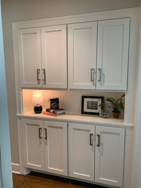 A built-in cabinet with white shaker-style doors and a softly lit display shelf holding books, a small plant, and framed decor | Dusty Rhodes.