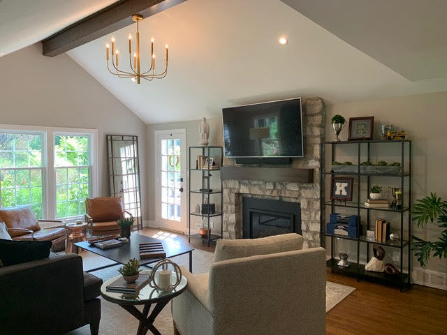 A vaulted living room with a stone fireplace, a mounted TV, and industrial-style shelving, complemented by comfortable seating and natural light | Dusty Rhodes.