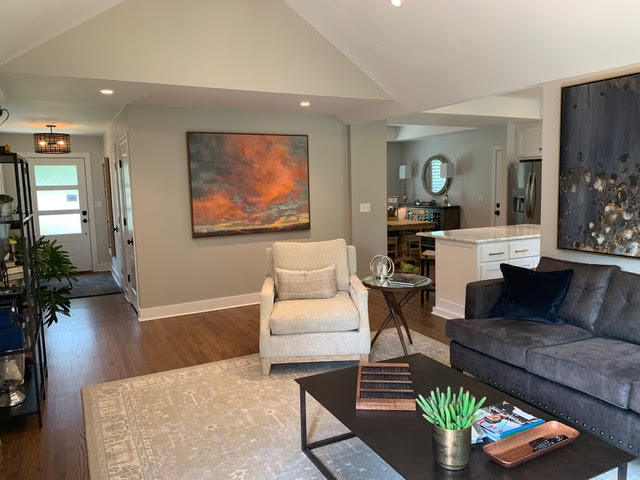 A living room featuring a gray sofa, a beige armchair, and a modern painting, with views into the kitchen and entryway | Dusty Rhodes.