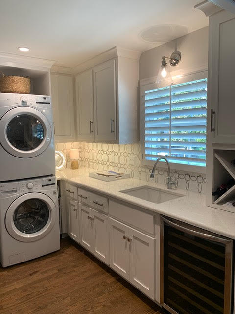 A modern laundry room with stacked washer and dryer, white cabinets, and a sleek countertop with a sink, illuminated by natural light through plantation shutters | Dusty Rhodes.